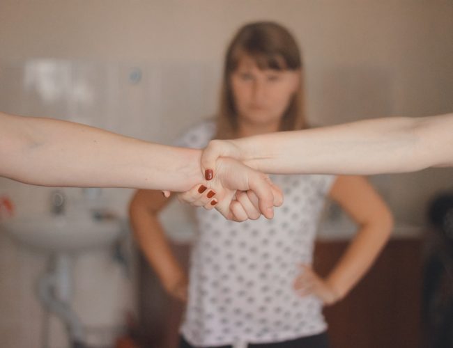 A self-defence trainer in a gym stands in front of two women clasping hands in a training session.