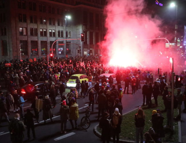 A street in Warsaw at night filled with women protesters with a pink flare and smoke in the background.
